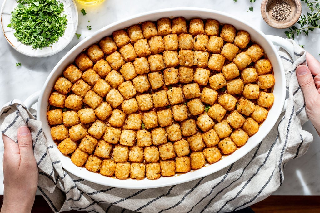 A white oval baking dish filled with neatly arranged, golden-brown tater tots sits on a striped cloth, with hands holding the sides. Chopped herbs, pepper, and oil are nearby on a white surface.