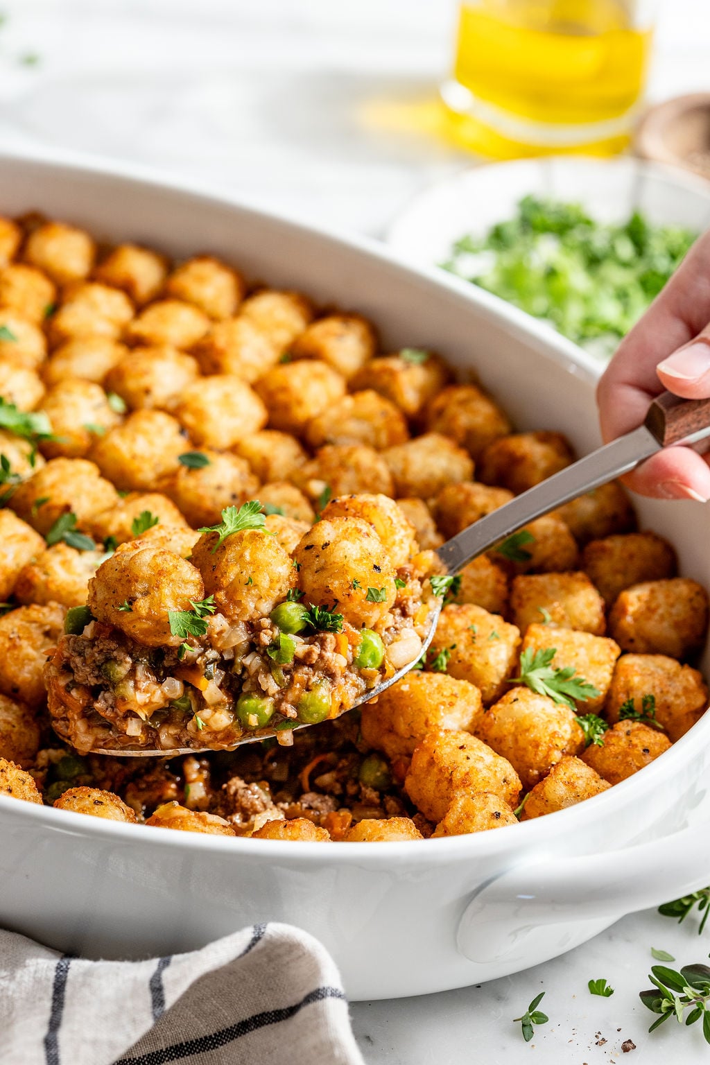 A hand lifts a serving of tater tot casserole from a white baking dish. The casserole is topped with golden tater tots and garnished with fresh herbs, with ground meat and vegetables visible inside.