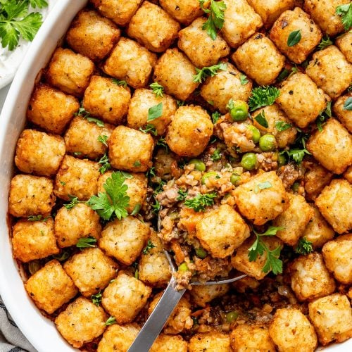 A baked casserole topped with crispy tater tots, garnished with fresh parsley. A serving spoon reveals a filling of vegetables and ground meat beneath the golden tater tot layer.