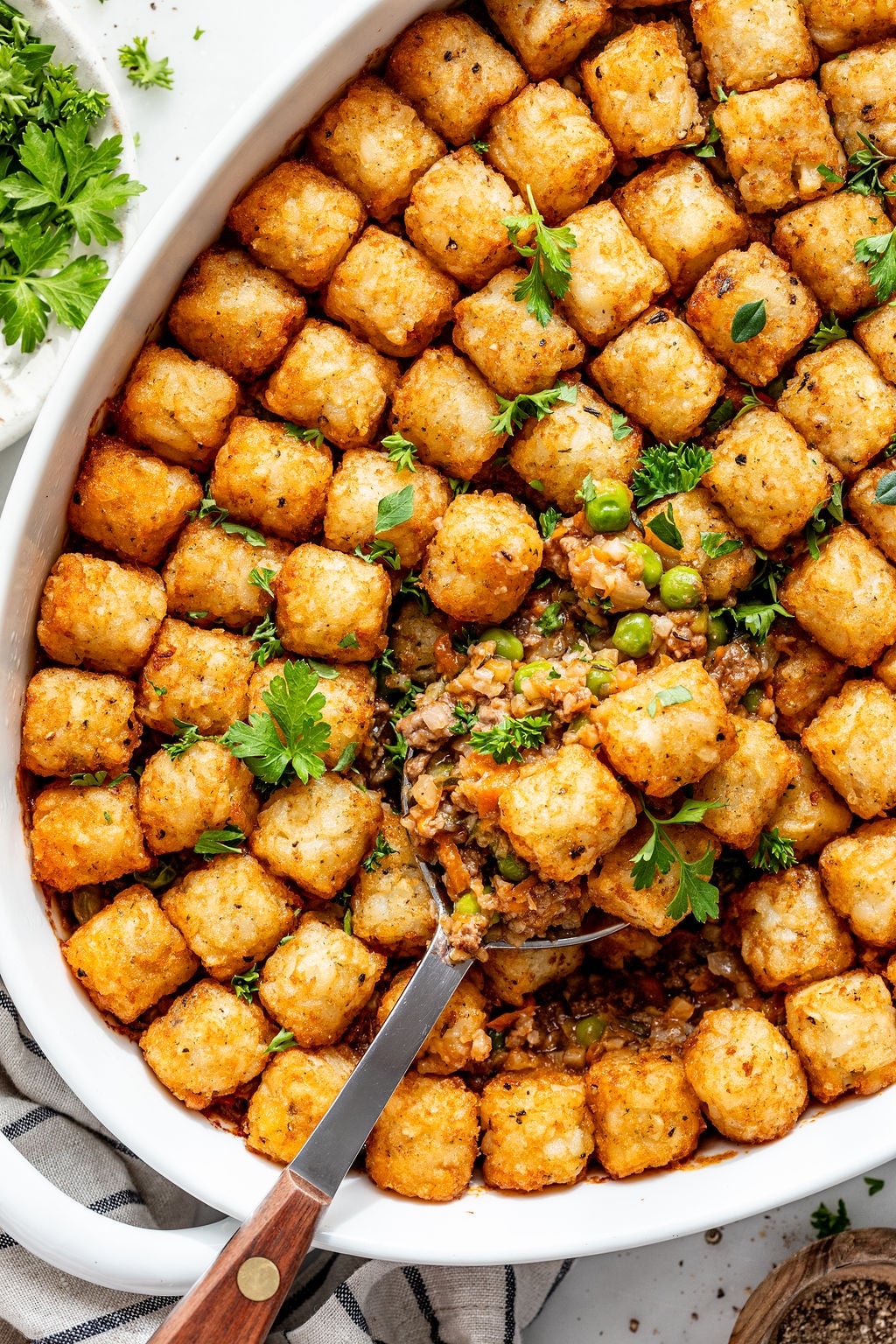 A baked casserole topped with crispy tater tots, garnished with fresh parsley. A serving spoon reveals a filling of vegetables and ground meat beneath the golden tater tot layer.