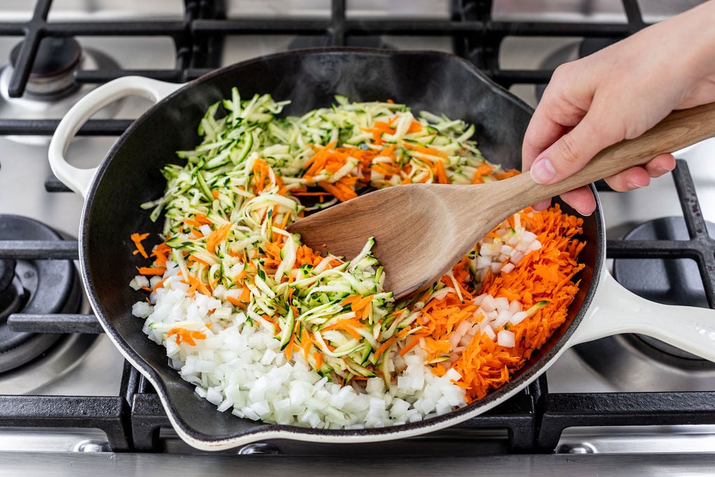 A hand stirs chopped onions, shredded carrots, and shredded zucchini in a white skillet on a gas stove.