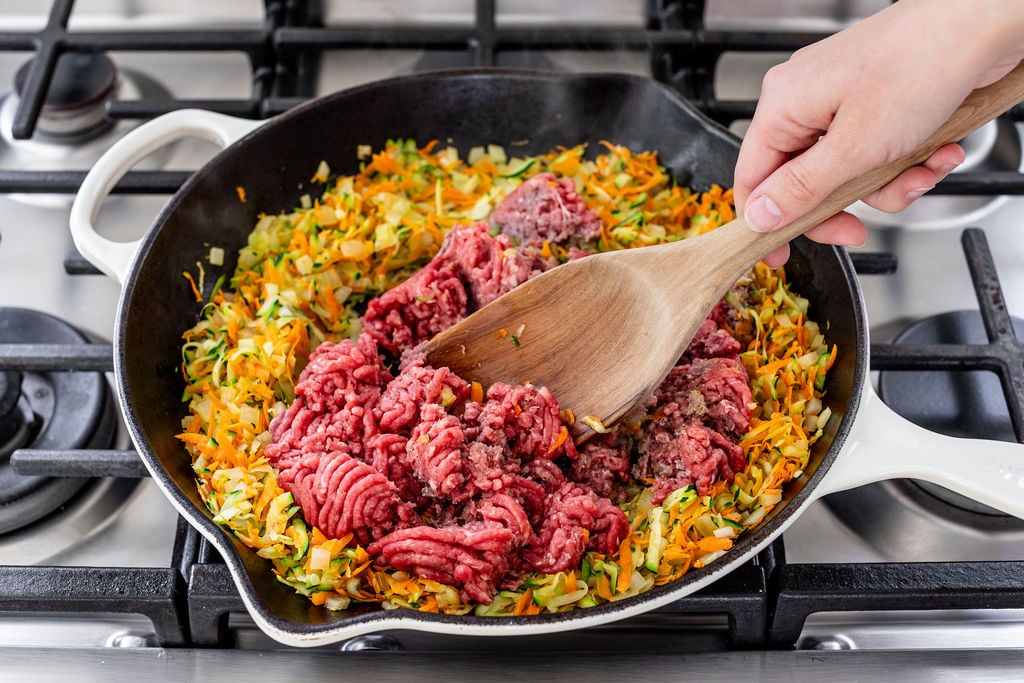 A hand uses a wooden spoon to sauté ground beef with chopped carrots, zucchini, and onions in a skillet on a stove.