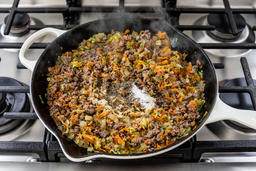 A skillet on a stove contains ground meat, chopped vegetables, and seasonings being cooked together. Steam rises from the mixture, and the ingredients are being mixed and sautéed in a white pan.