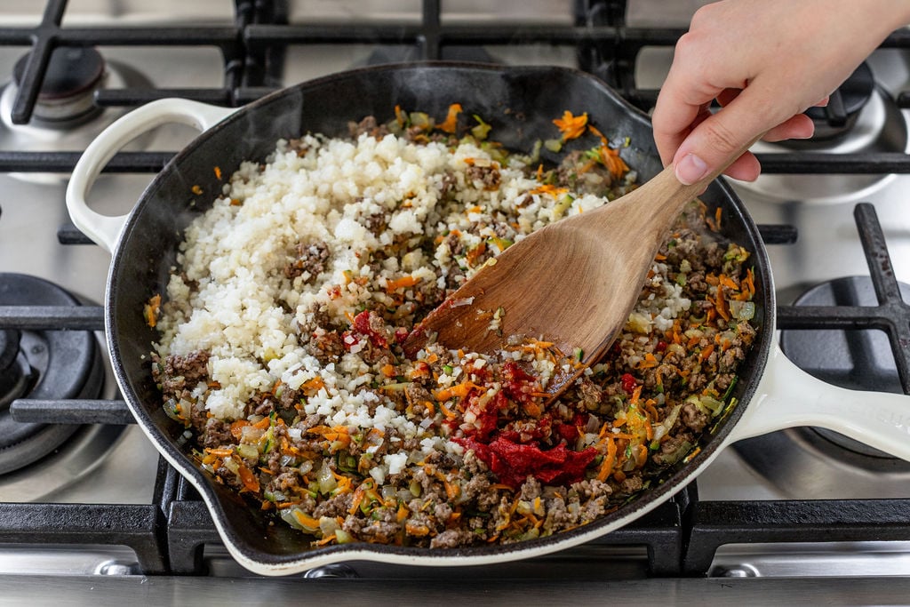 A hand stirs a mixture of ground meat, vegetables, tomato paste, and cauliflower rice in a white skillet on a gas stove.