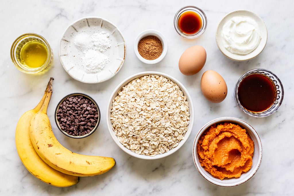 Oats, chocolate chips, bananas, pumpkin puree, eggs, oil, yogurt, maple syrup, vanilla, baking powder, and cinnamon in bowls on a white marble surface—all the ingredients you need to make delicious banana pumpkin muffins.