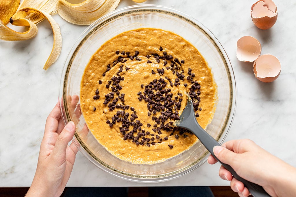 A person stirs chocolate chips into banana bread batter in a glass bowl, preparing it much like they would for banana pumpkin muffins. Eggshells and banana peels are on a white marble countertop nearby.