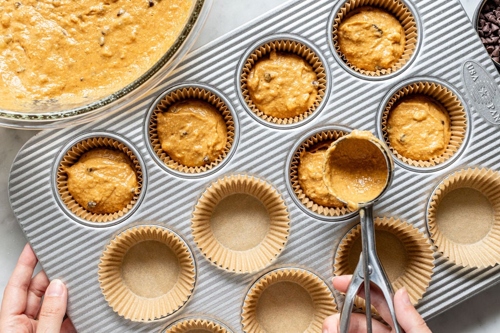 A person fills muffin liners in a baking tray with batter for banana pumpkin muffins using an ice cream scoop. Some liners are filled, while others remain empty. A glass bowl with more banana pumpkin muffin batter is nearby.