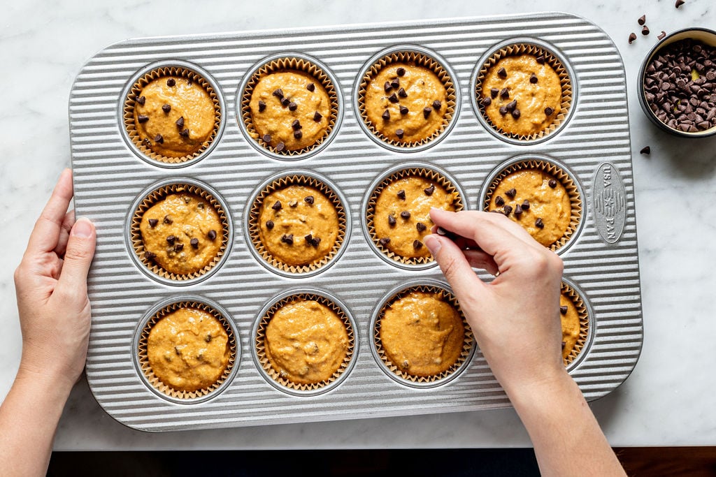 A person adds chocolate chips to banana pumpkin muffin batter in a muffin tray, which sits on a marble countertop. A small bowl of extra chocolate chips is nearby.