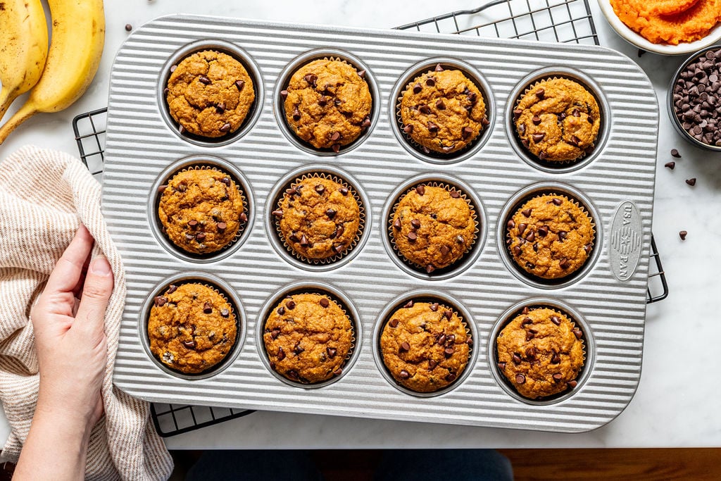A person holds a muffin pan filled with twelve freshly baked banana pumpkin muffins topped with chocolate chips, resting on a cooling rack. Bananas, pumpkin puree, and chocolate chips are nearby.