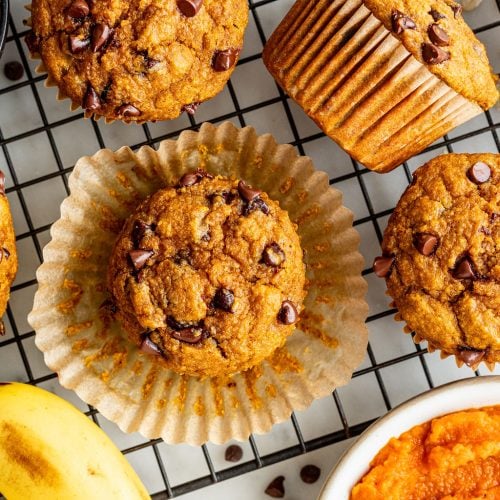 Banana pumpkin muffins with chocolate chips rest on a cooling rack, surrounded by a bowl of pumpkin puree, a ripe banana, oats, and extra chocolate chips.