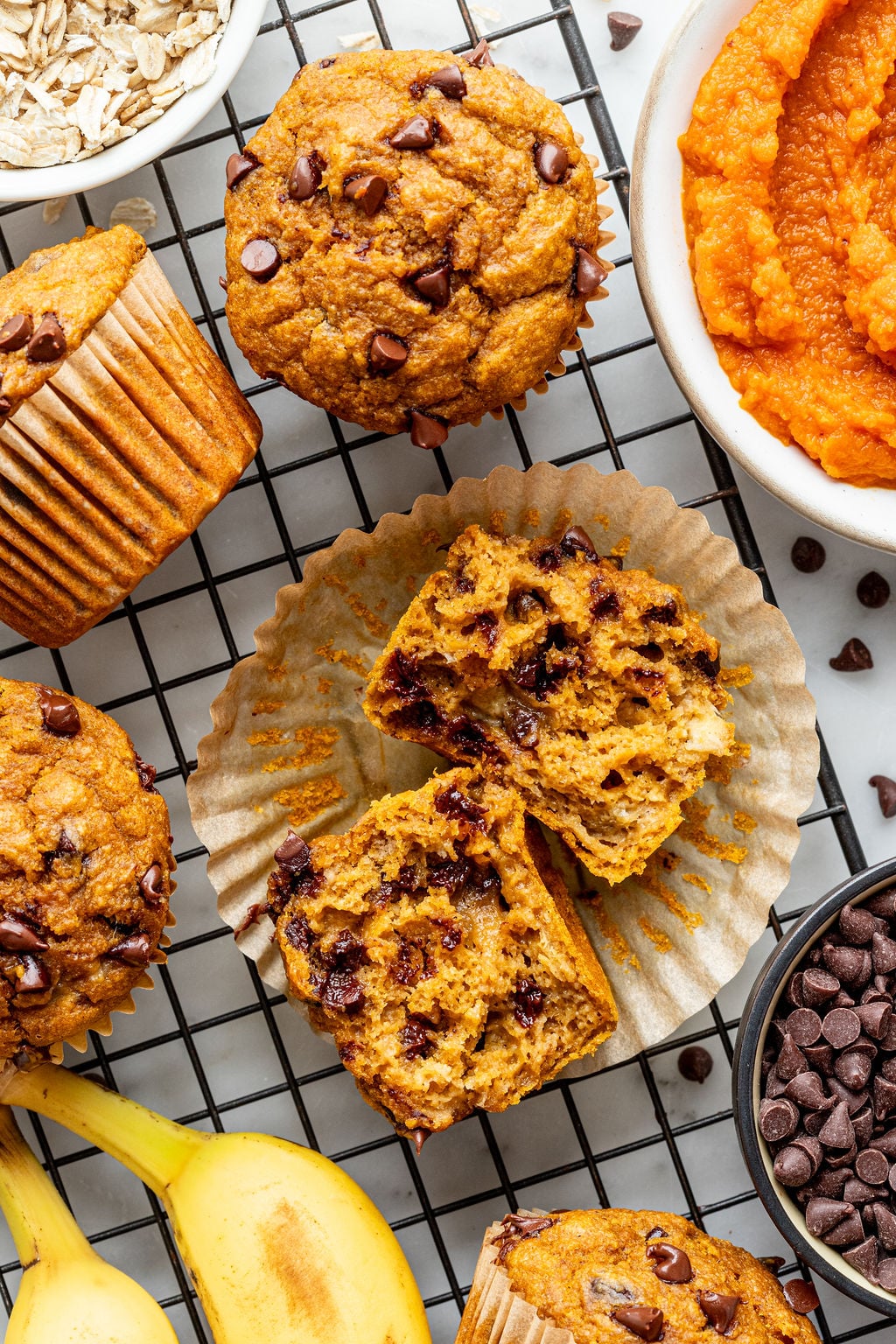 Banana pumpkin muffins with chocolate chips rest on a cooling rack, with one muffin cut in half to reveal its moist interior. They’re surrounded by a bowl of oats, mashed pumpkin, chocolate chips, and ripe bananas—the perfect ingredients for these irresistible treats.