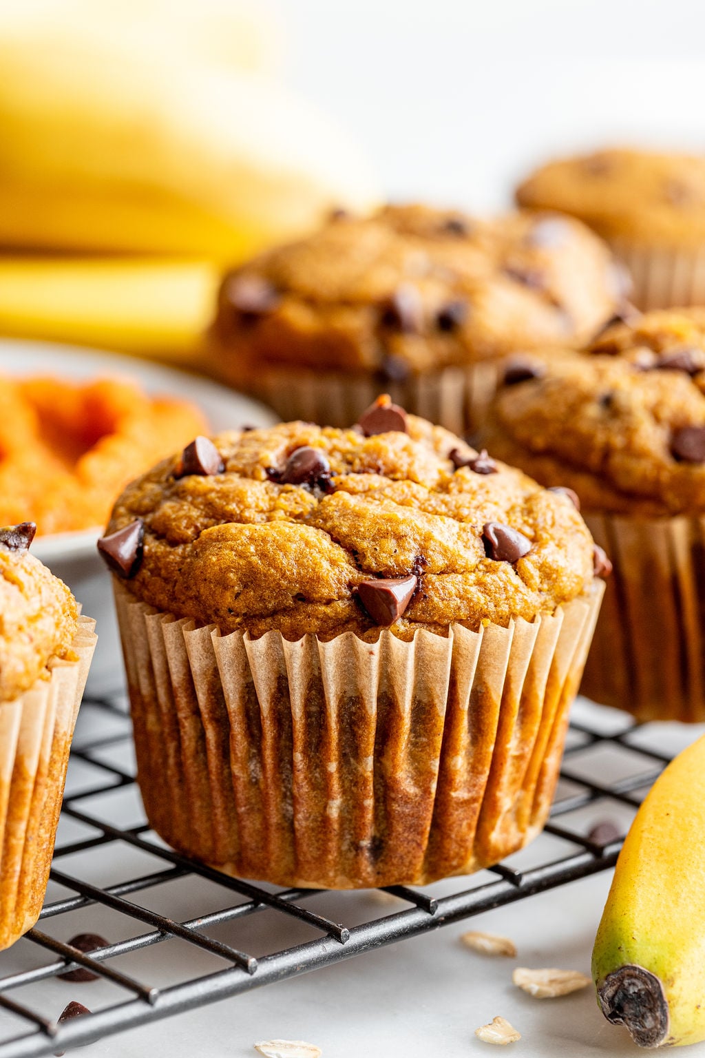 A close-up of banana pumpkin muffins cooling on a wire rack, with chocolate chips, bananas, and oats visible nearby.