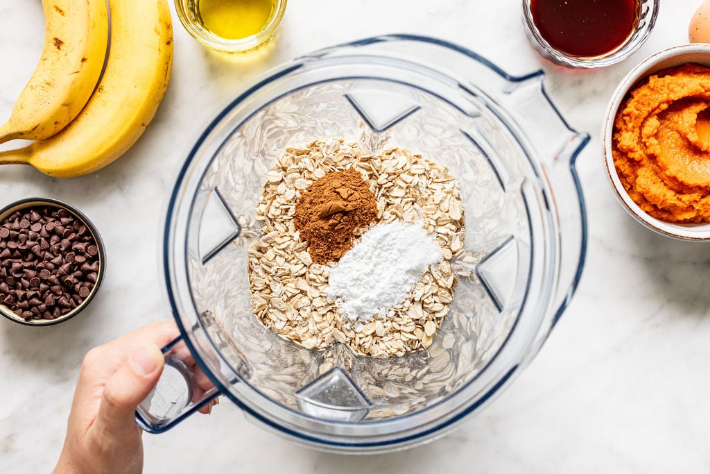 A blender with oats, cinnamon, and baking powder inside, surrounded by bananas, chocolate chips, olive oil, a bowl of pumpkin puree, a bowl of dark liquid, and an egg on a white surface—everything you need to whip up delicious banana pumpkin muffins.