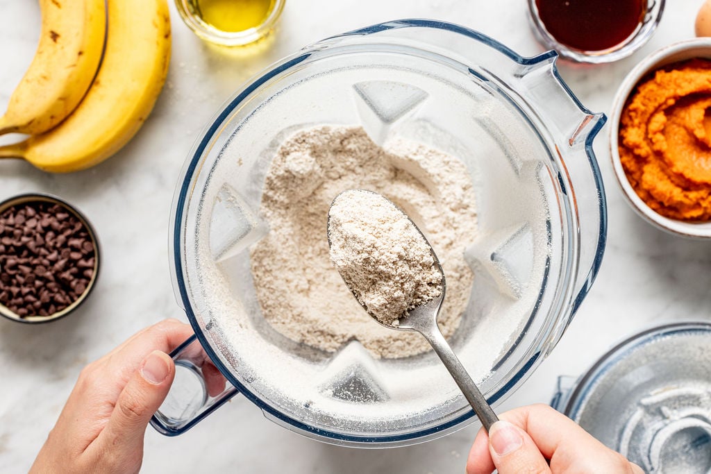 A hand holds a spoon with flour over a blender, preparing the batter for delicious banana pumpkin muffins. Surrounding the blender are bananas, a bowl of chocolate chips, a bowl of pumpkin puree, oil, and a red liquid in a bowl.