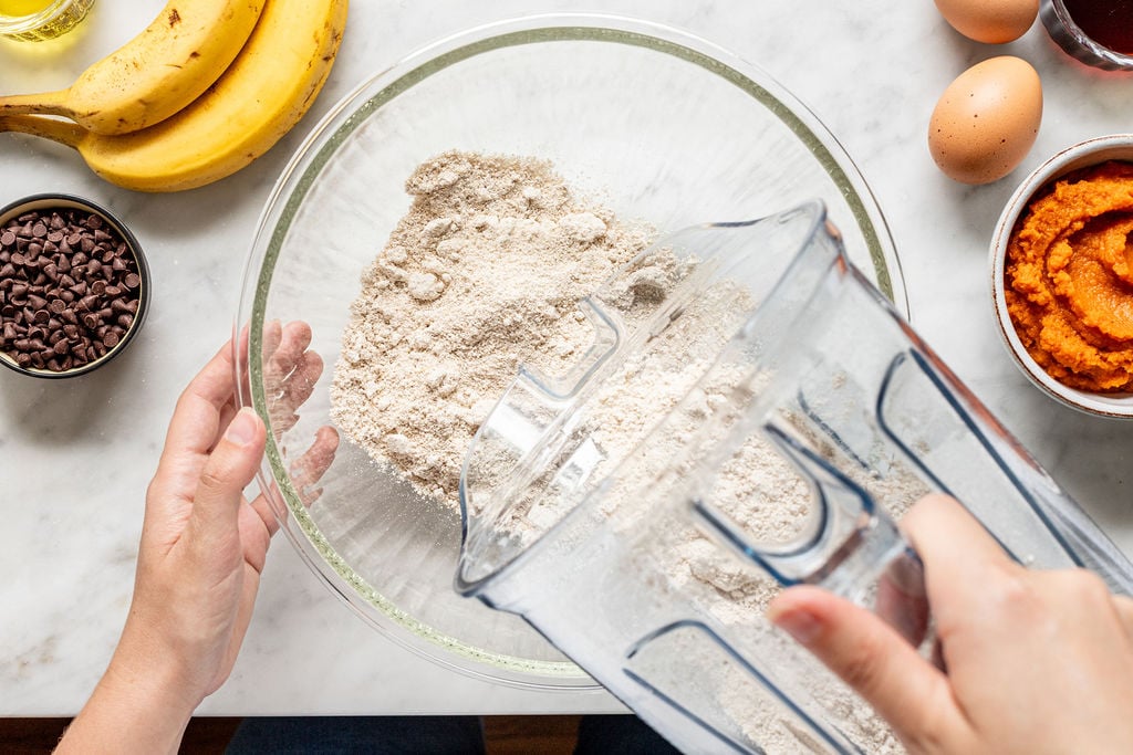A person pours flour from a blender into a glass bowl. Surrounding the bowl are ingredients for banana pumpkin muffins, including ripe bananas, eggs, chocolate chips, a bowl of pumpkin puree, and other baking essentials on a marble countertop.