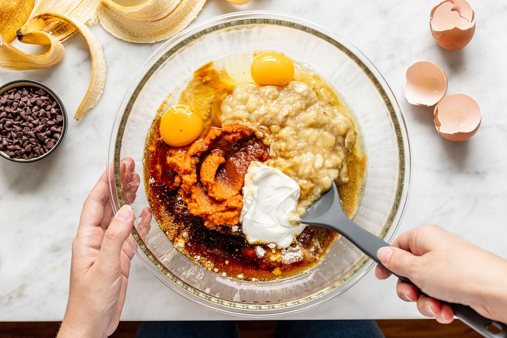 Hands holding a bowl with eggs, mashed banana, pumpkin puree, yogurt, and spices being mixed for banana pumpkin muffins; eggshells, banana peel, and chocolate chips are nearby on a marble counter.