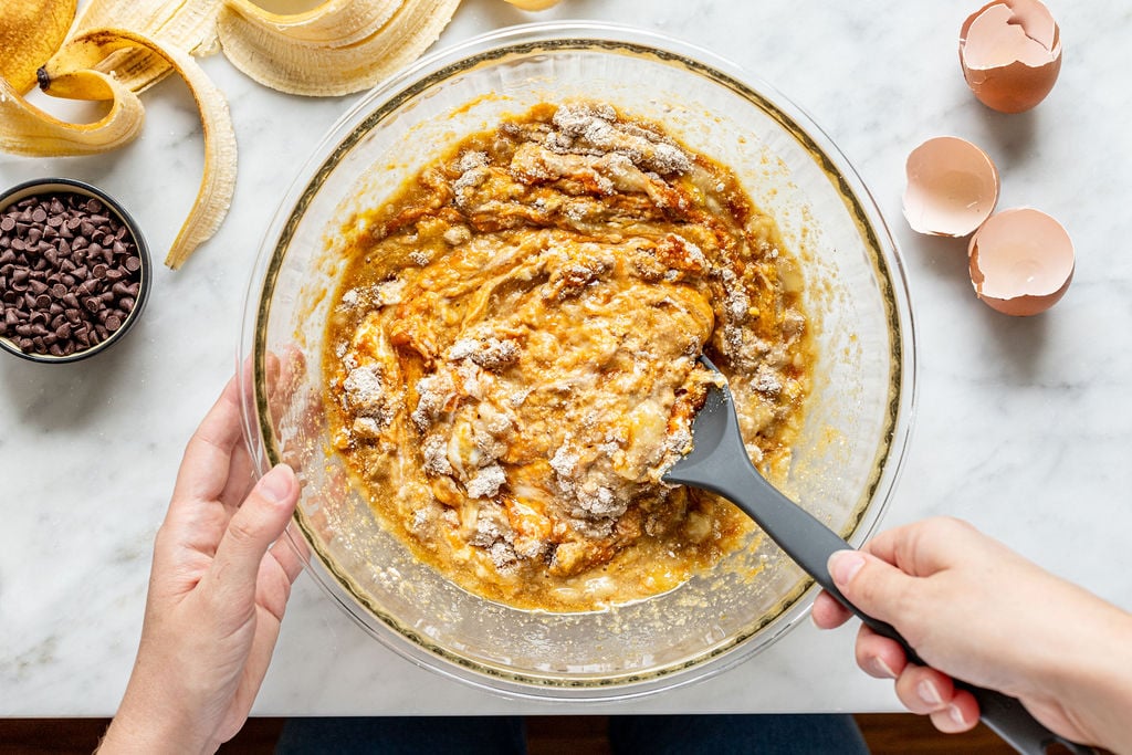 A person mixing banana bread batter in a glass bowl with a spatula, perhaps inspired to try making banana pumpkin muffins next. Nearby are banana peels, chocolate chips, and eggshells scattered across the marble countertop.