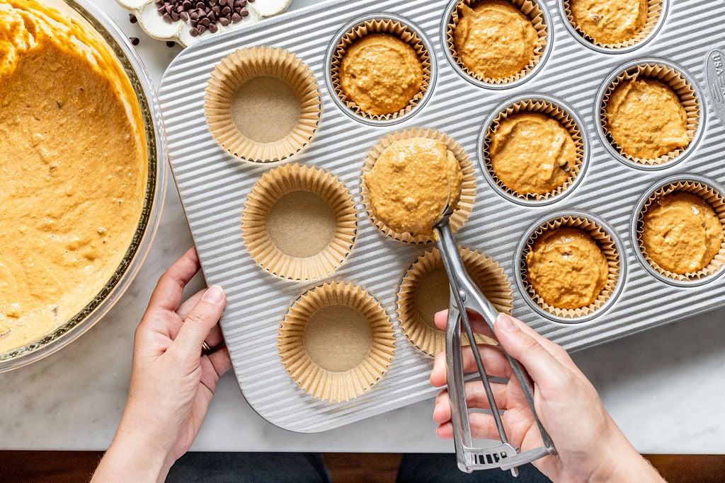 A person uses an ice cream scoop to fill muffin liners with batter for cottage cheese pumpkin muffins in a muffin tin. A bowl of batter and chocolate chips are nearby on a white countertop.