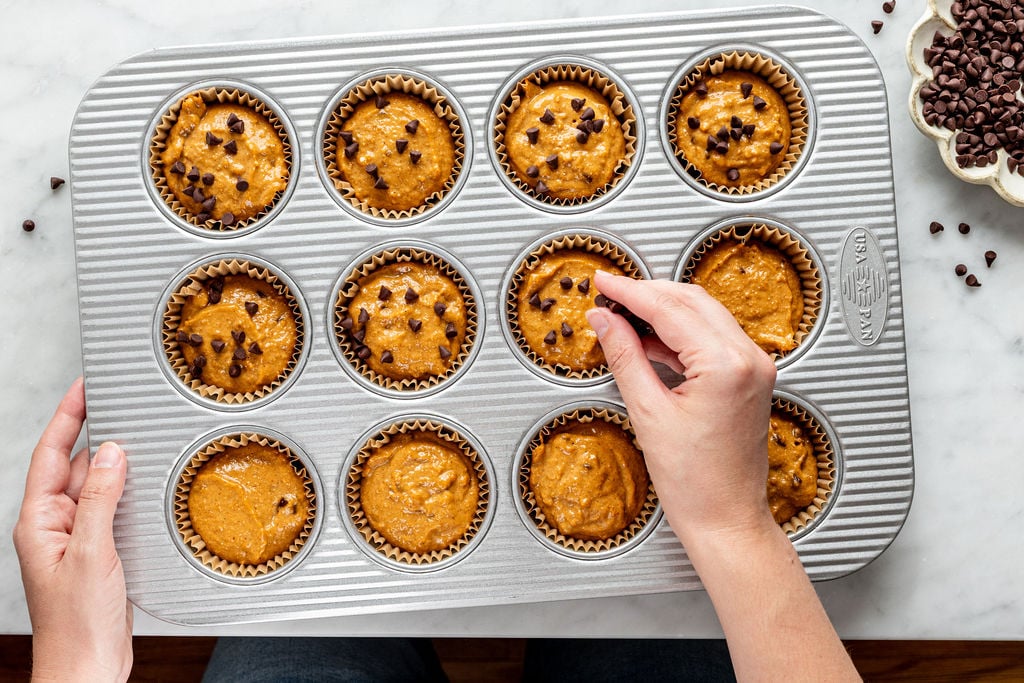 A person places chocolate chips on cottage cheese pumpkin muffin batter in a 12-cup muffin tin lined with paper liners. A small bowl of chocolate chips sits nearby on a marble surface.