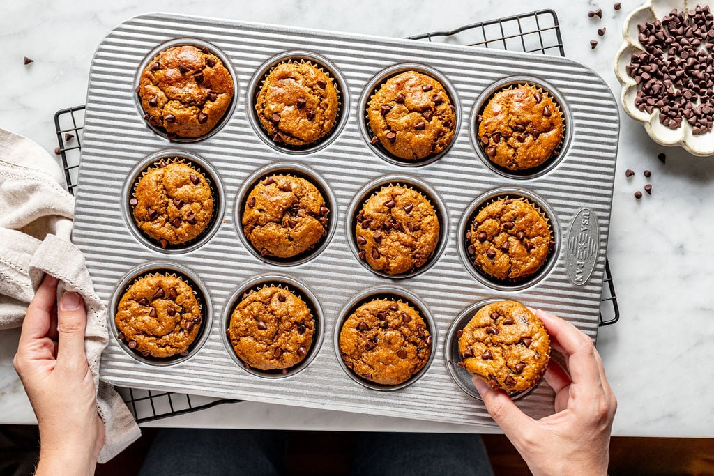 A person holds a muffin tin filled with freshly baked cottage cheese pumpkin muffins, with one muffin being lifted out. A bowl of chocolate chips and a cooling rack are nearby.