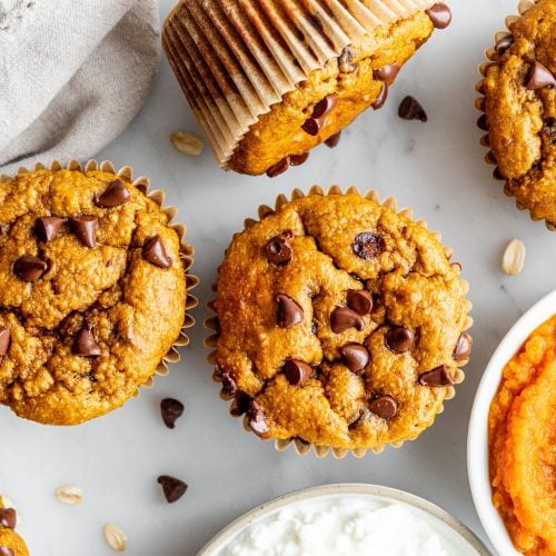 Pumpkin chocolate chip muffins arranged on a white surface, surrounded by a bowl of pumpkin puree, a bowl of cottage cheese, scattered chocolate chips, and a beige napkin.