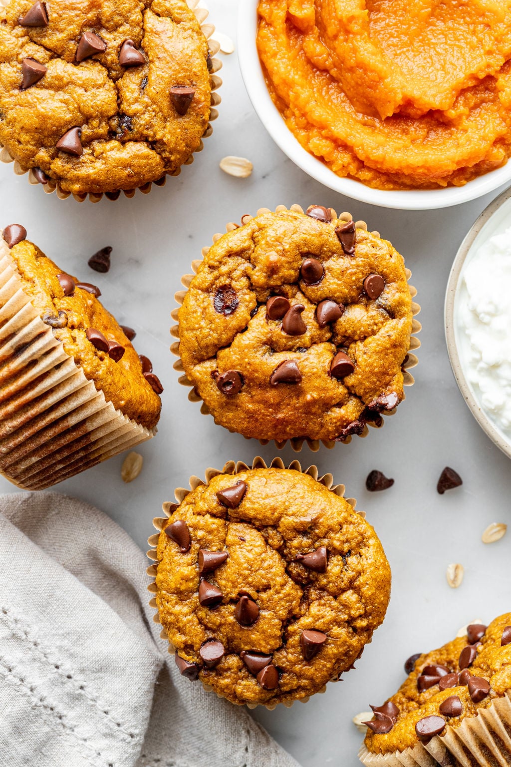Pumpkin chocolate chip muffins arranged on a white surface, surrounded by a bowl of pumpkin puree, a bowl of cottage cheese, scattered chocolate chips, and a beige napkin.