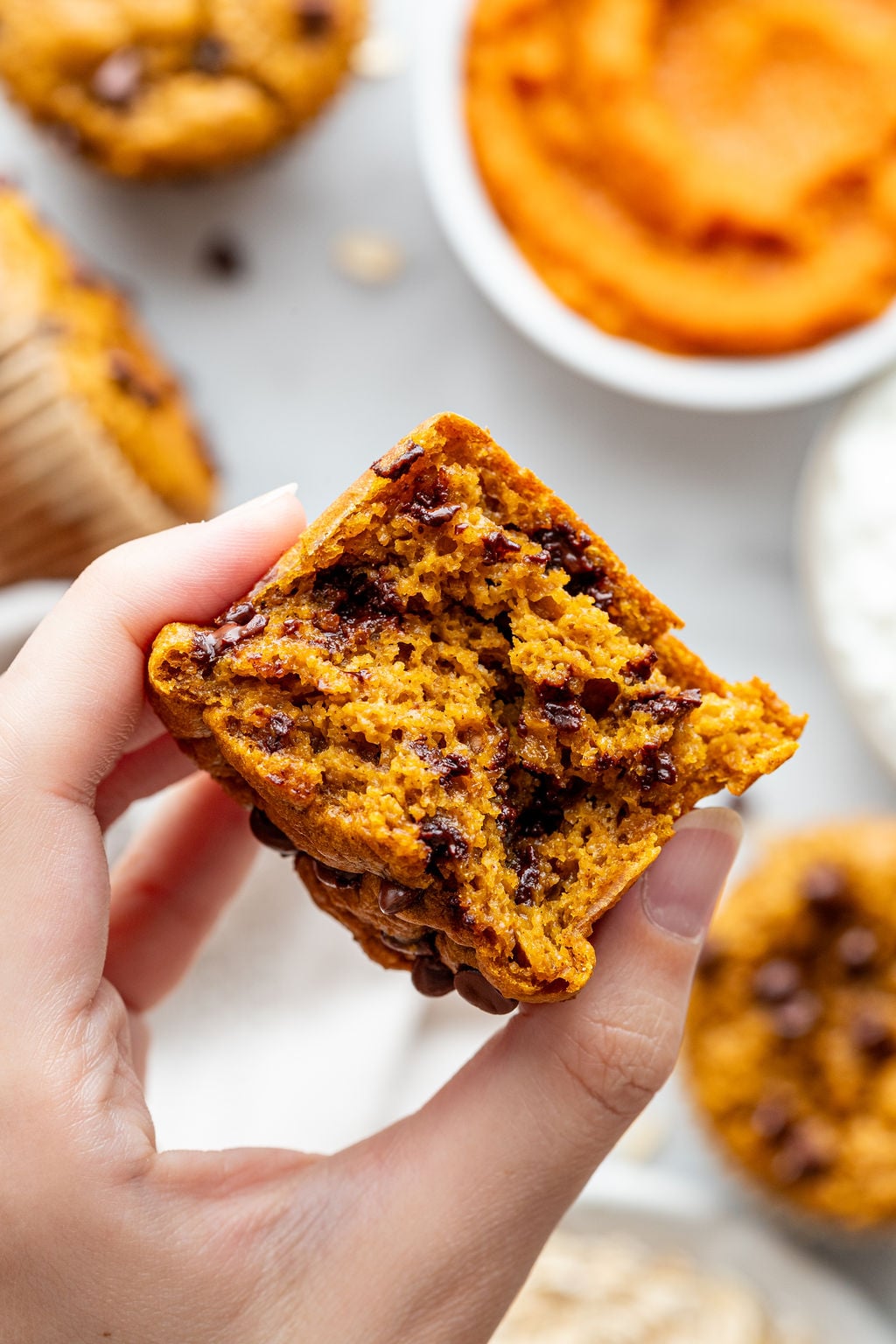 A hand holds a cottage cheese pumpkin muffin with chocolate chips, broken in half to reveal the moist, orange interior. Other muffins and a bowl of pumpkin puree are visible in the blurred background.