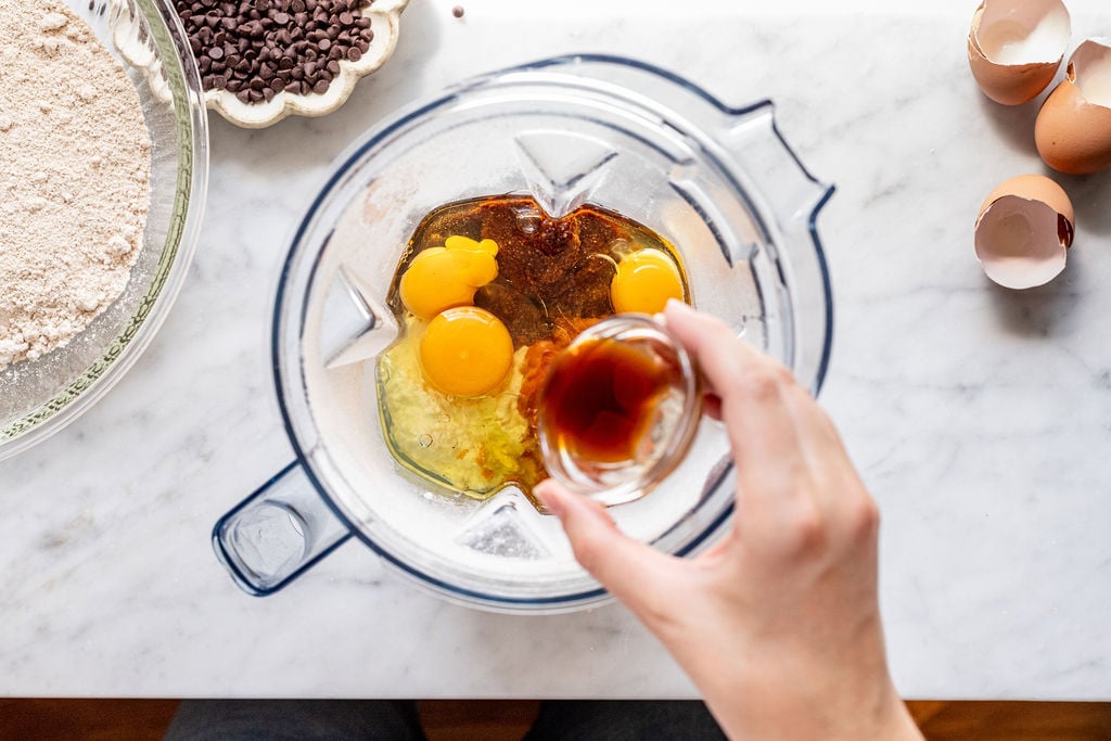 A hand pours vanilla extract into a blender containing eggs, wet ingredients, and creamy cottage cheese for pumpkin muffins, with a bowl of flour, chocolate chips, and cracked eggshells visible on a marble countertop.