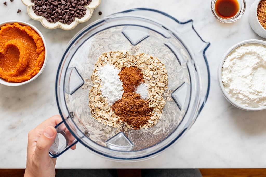 A hand holds a blender filled with oats, baking powder, salt, and cinnamon. Surrounding bowls hold pumpkin puree, chocolate chips, vanilla extract, and flour on a white countertop.