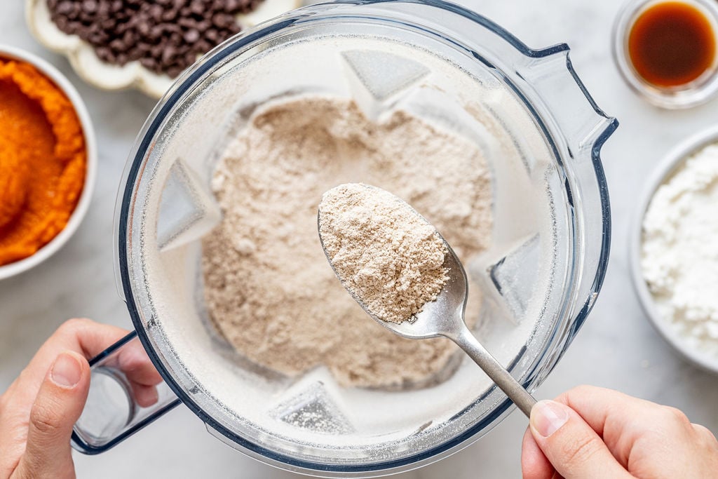 A hand holds a spoonful of flour over a blender filled with flour. Surrounding bowls contain chocolate chips, pumpkin puree, vanilla extract, and more flour on a white surface.