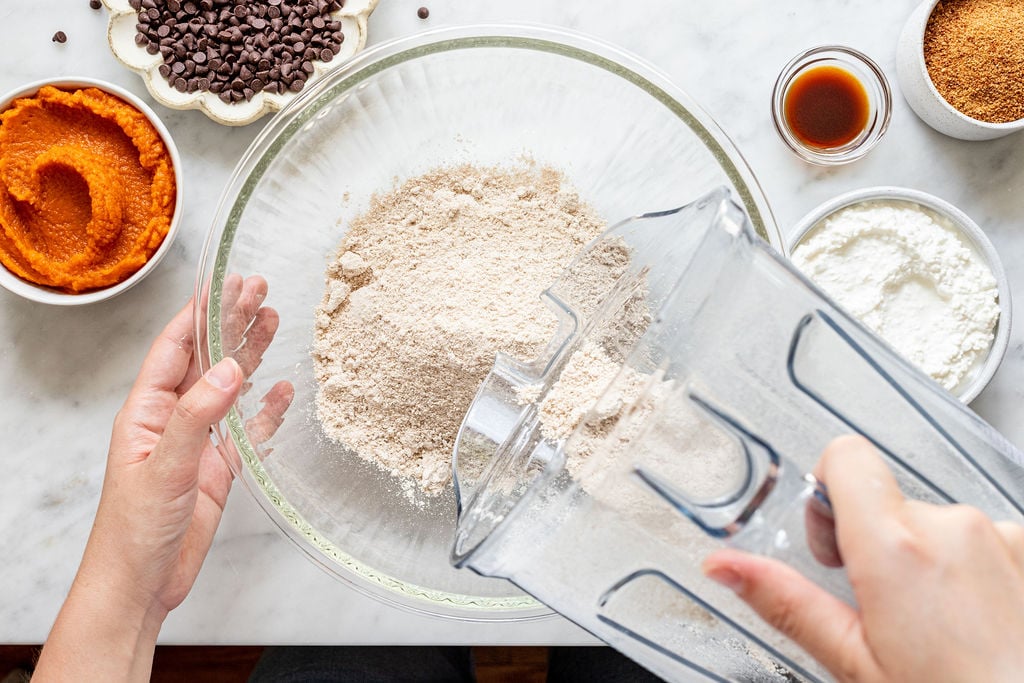 A person pours flour from a blender into a glass bowl surrounded by chocolate chips, pumpkin puree, brown sugar, vanilla, and more flour on a marble countertop.