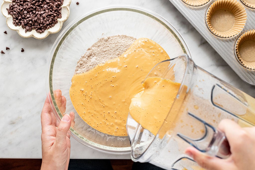 A person pours a blended wet mixture into a bowl of dry ingredients on a marble counter, preparing to make cottage cheese pumpkin muffins. Chocolate chips and a muffin tin with paper liners are nearby.