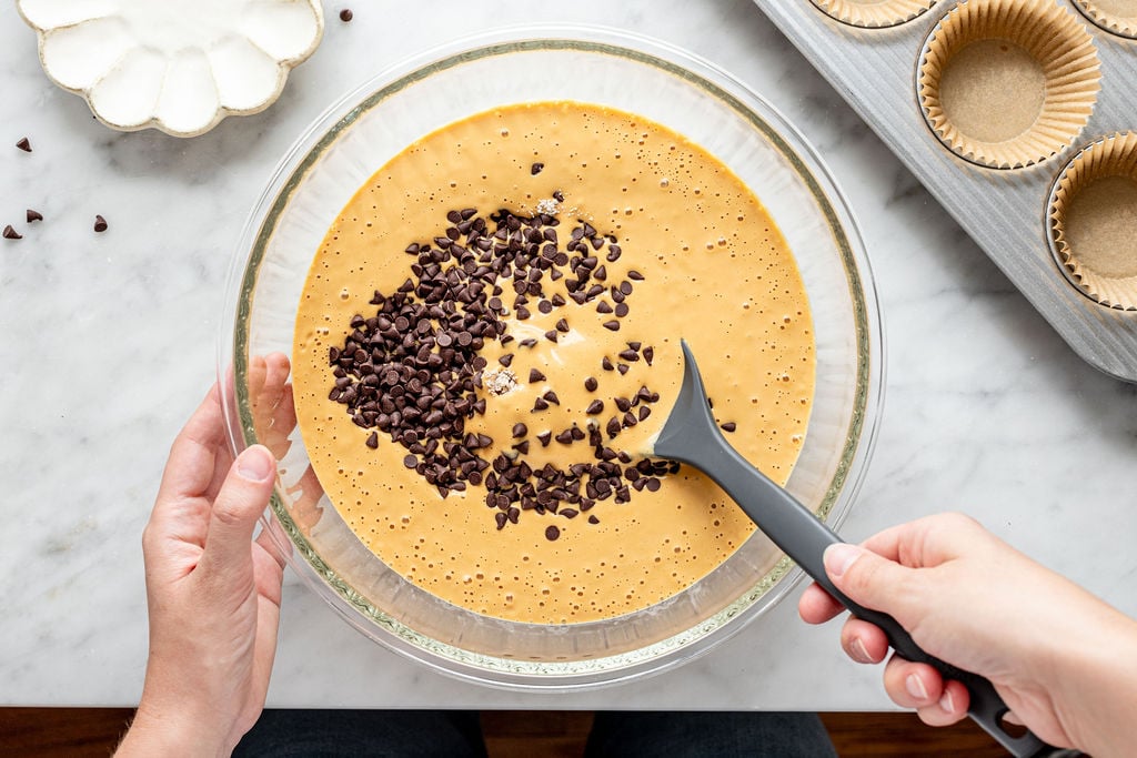 A person stirs chocolate chips into a bowl of yellow batter with a black spatula, preparing delicious cottage cheese pumpkin muffins. Cupcake liners and a white dish with more chocolate chips are nearby on a marble countertop.
