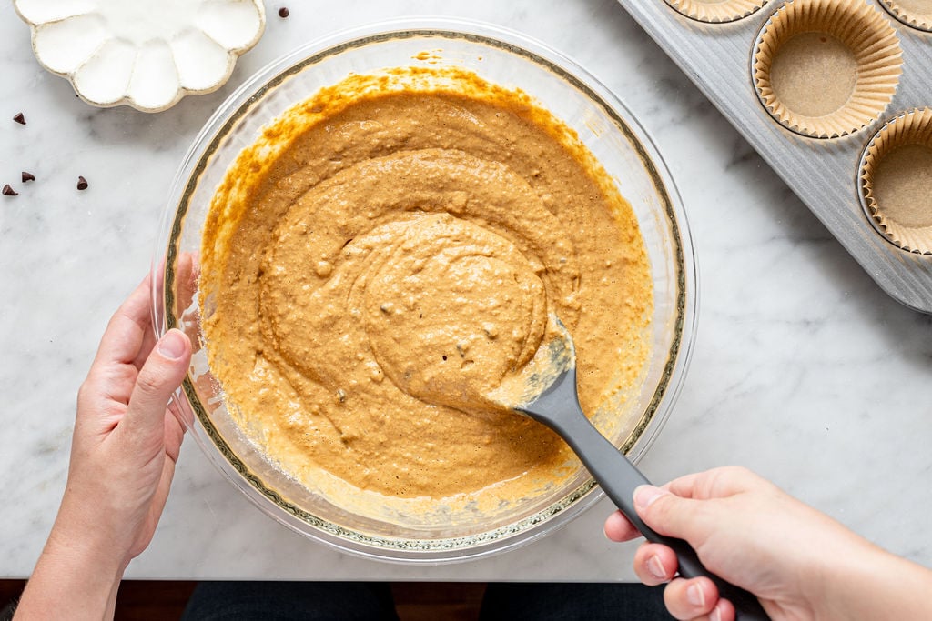 A person stirs cottage cheese pumpkin muffin batter in a glass bowl with a spatula. Cupcake liners are arranged in a baking tray nearby on a marble countertop.