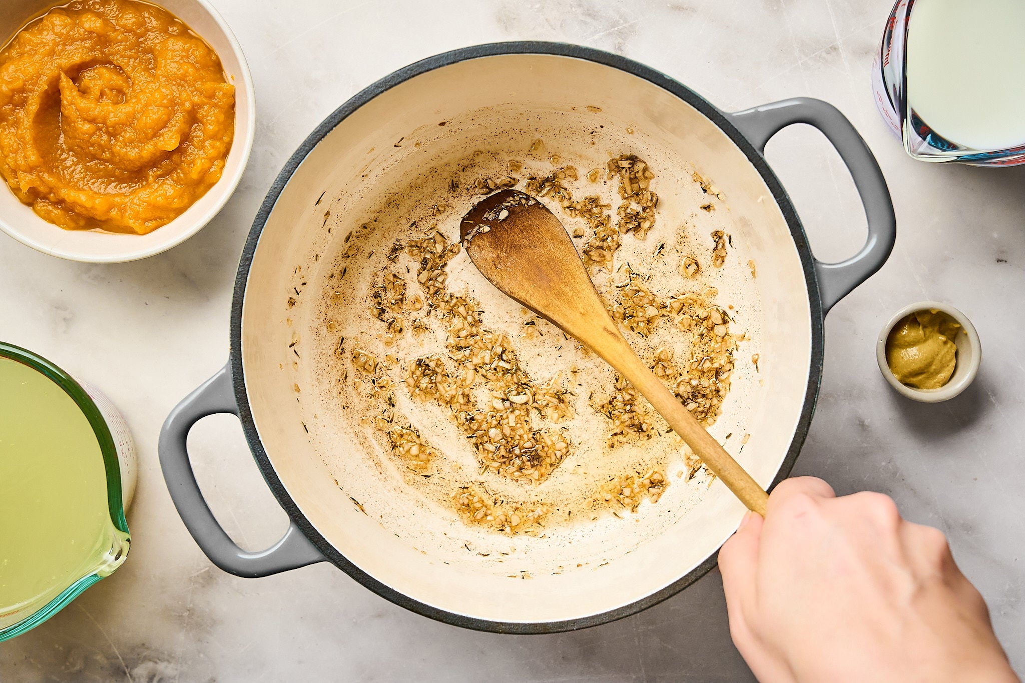 A hand stirs sautéed onions and garlic in a large pot with a wooden spoon, the first step in crafting a cozy pumpkin mac and cheese. Surrounding the pot are bowls of broth, pumpkin purée, and a small dish of mustard.