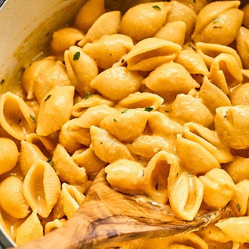 A wooden spoon stirs creamy, orange-colored shell pasta in a large pot, with luscious pumpkin mac and cheese sauce clinging to the noodles.