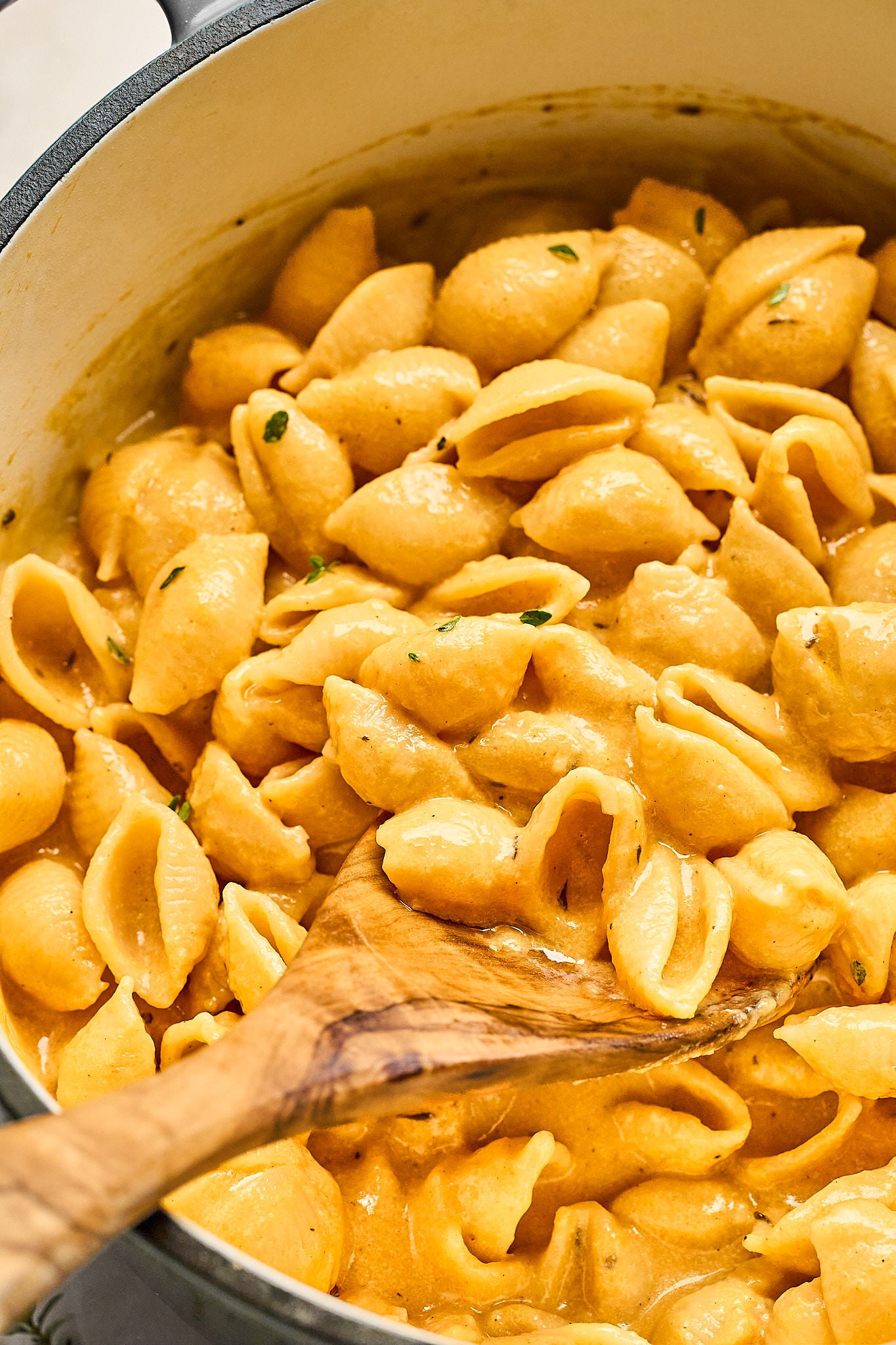 A wooden spoon stirs creamy, orange-colored shell pasta in a large pot, with luscious pumpkin mac and cheese sauce clinging to the noodles.
