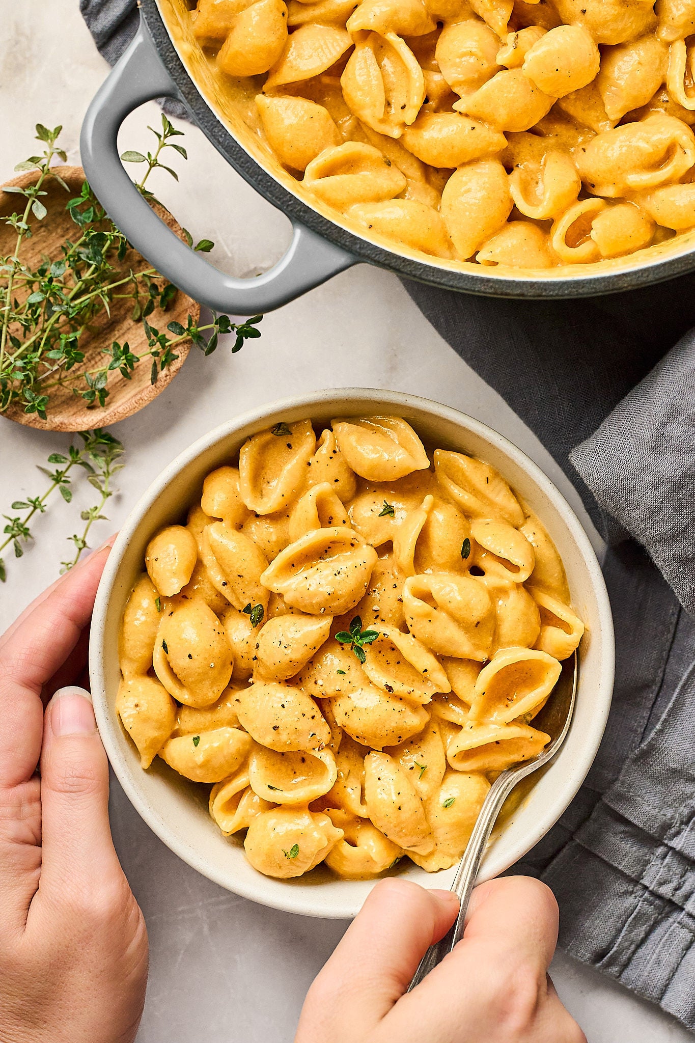 A bowl of creamy pumpkin mac and cheese shell pasta topped with black pepper and fresh thyme, held by one hand with a fork in the other. A pot of more pasta and a small dish of thyme are nearby.
