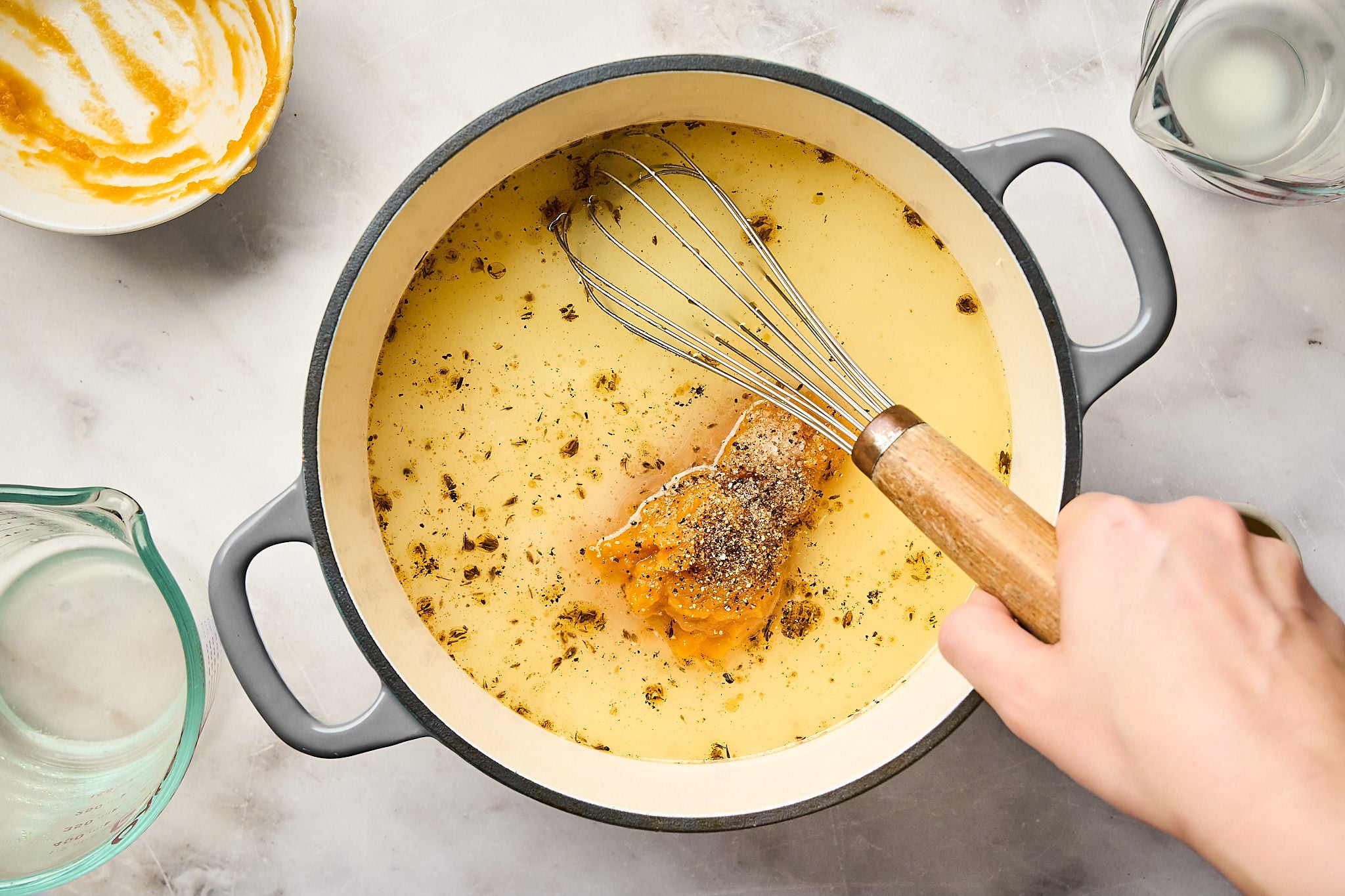 A hand whisks broth with herbs and seasonings in a large gray pot, preparing a soup or sauce—perhaps the base for a creamy pumpkin mac and cheese. Mixing bowls and a measuring cup are nearby on a light countertop.