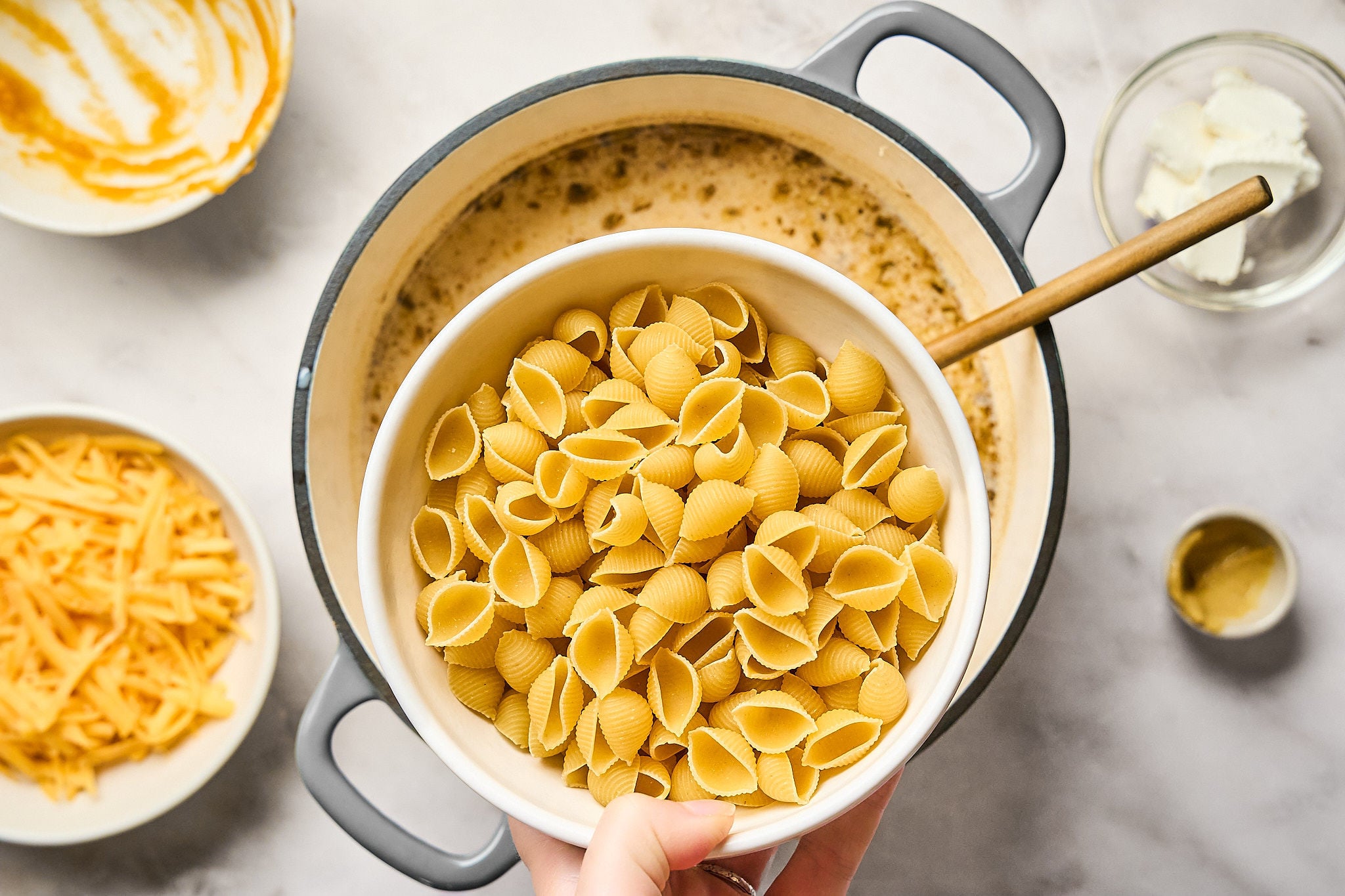 A hand holds a bowl of uncooked shell pasta over a pot of simmering liquid, surrounded by bowls of shredded cheese, cream cheese, and mustard on a marble countertop—ingredients neatly arranged for homemade pumpkin mac and cheese.