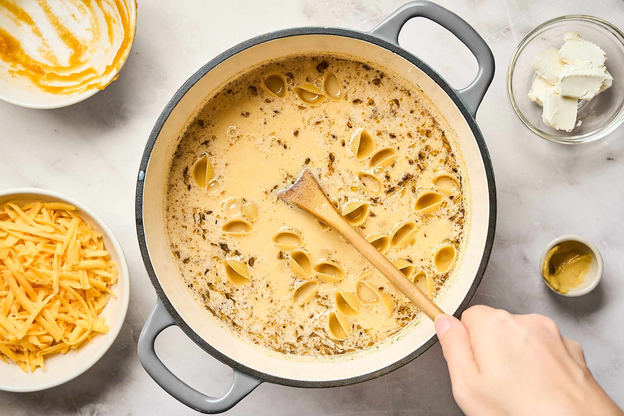 A hand stirs pasta shells in a creamy, seasoned sauce in a pot, making a comforting pumpkin mac and cheese. Surrounding the pot are bowls of shredded cheese, cream cheese, mustard, and a mixing bowl with sauce residue.