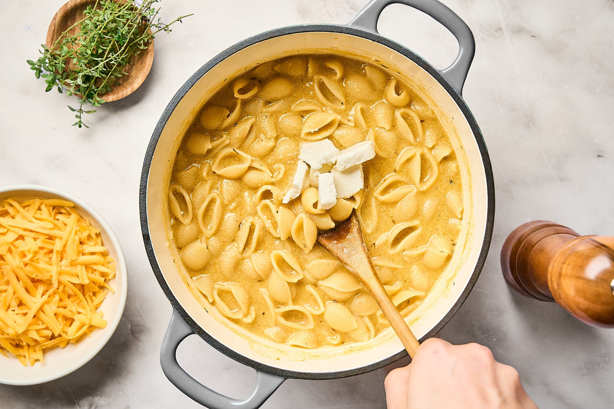 A hand stirs a pot of shell pasta with melted cheese, cream cheese cubes, and velvety pumpkin mac and cheese sauce. Shredded cheddar, fresh herbs, and a pepper grinder are nearby on a marble countertop.