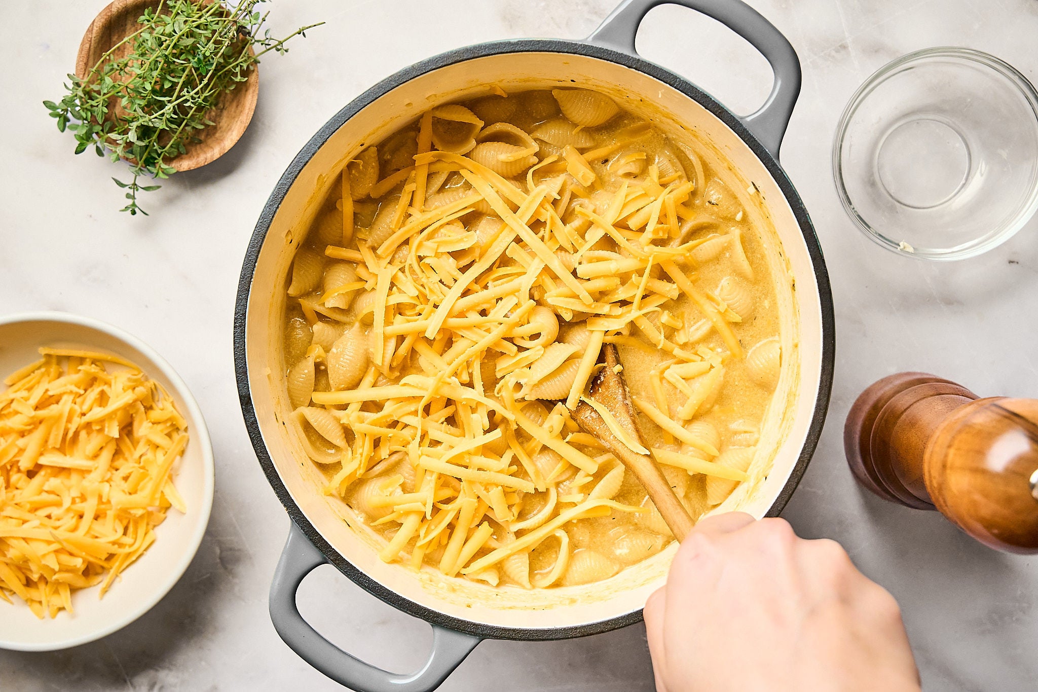 A hand stirs a pot of creamy pumpkin mac and cheese with shredded cheddar on top. Nearby are a bowl of cheese, fresh herbs, and a pepper grinder on a marble surface.