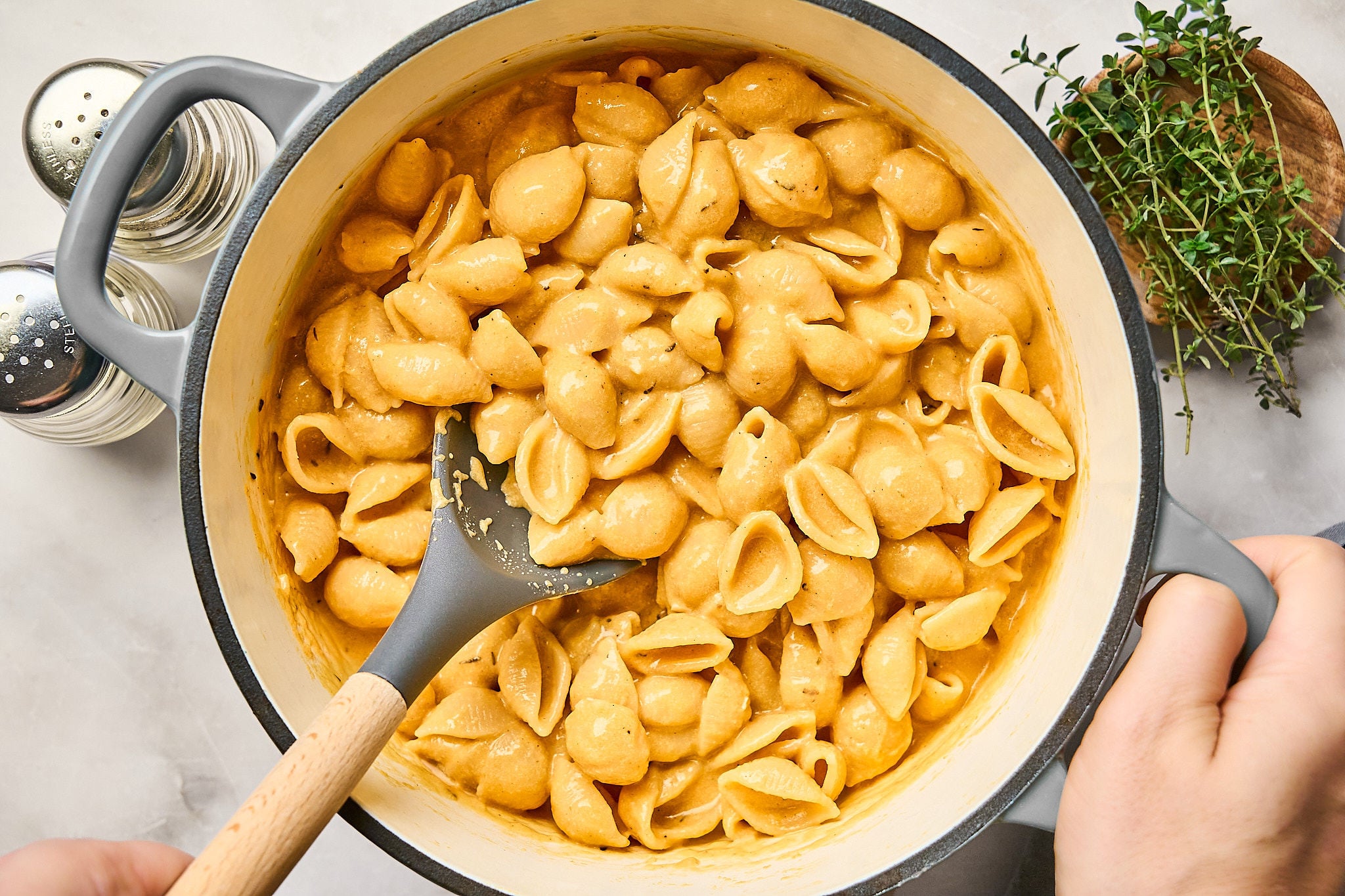 A pot of creamy pumpkin mac and cheese shell pasta with a black spoon, salt and pepper shakers, and fresh thyme on the side. Two hands are holding the pot on a light countertop.